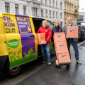 Three people standing beside a colorful delivery van on a city street, holding orange boxes labeled "Veloce." The van features the text "BILLA Online Shop." The scene suggests a delivery or logistics setting.