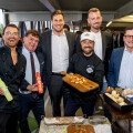 A group of six men smiling together at an event. Four of them are dressed in formal suits, while two wear chef attire. They are gathered around a table with appetizers and various condiments. One chef holds a tray with small servings of food. The setting appears to be a well-lit indoor venue with other attendees in the background.