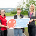 Three people standing outdoors holding a sign with a red smiley face and the text "Aufrunder bewirken Wunder." They are smiling, with greenery and a road in the background.