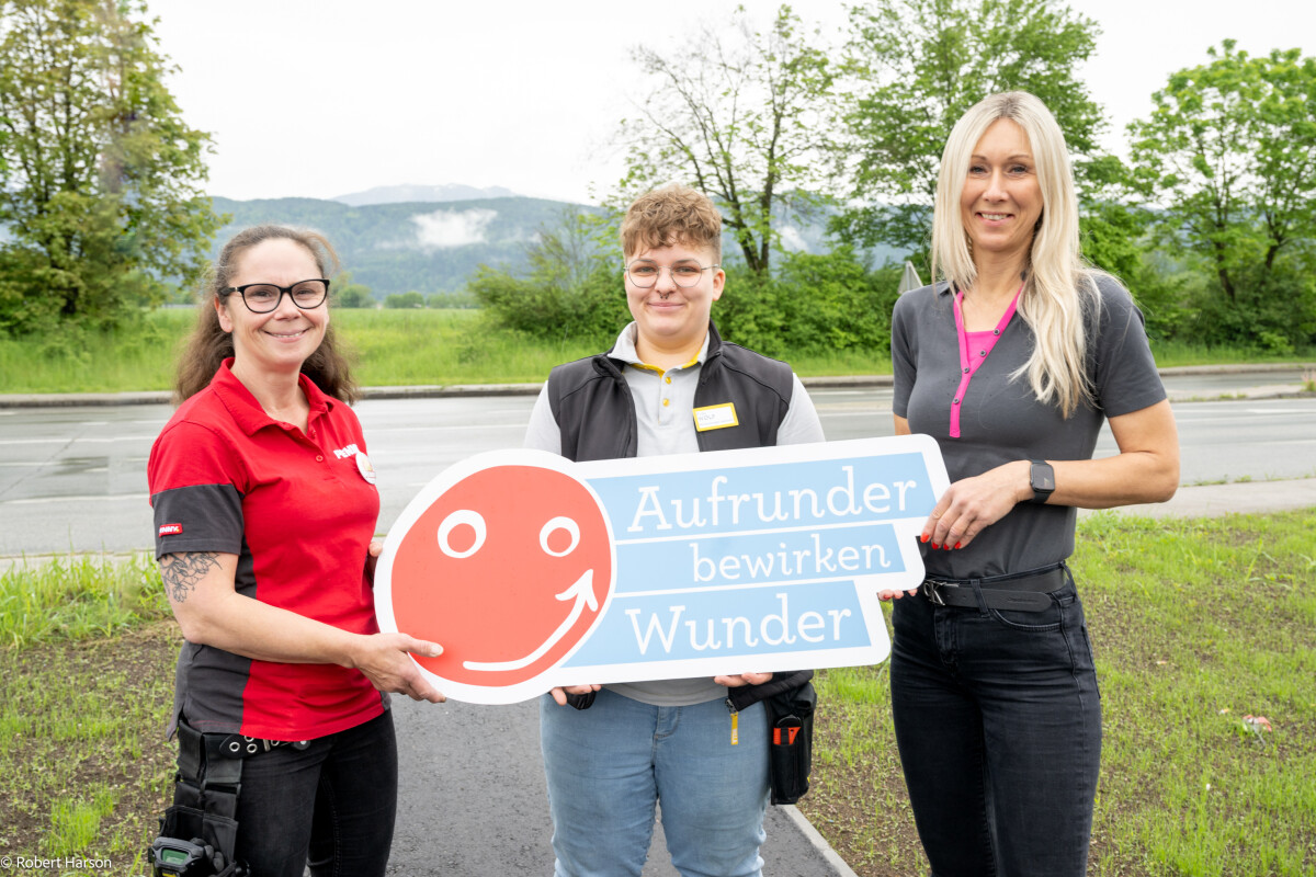 Three people outdoors holding a sign with a smiling face and the text "Aufrunder bewirken Wunder." They are standing on a grassy area with trees and a road in the background. One person is wearing a red shirt, another a dark vest, and the third a gray polo shirt.