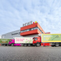 Three colorful trucks parked in front of a REWE distribution center, under a cloudy sky. The trucks display various German texts promoting green energy and electric transport.