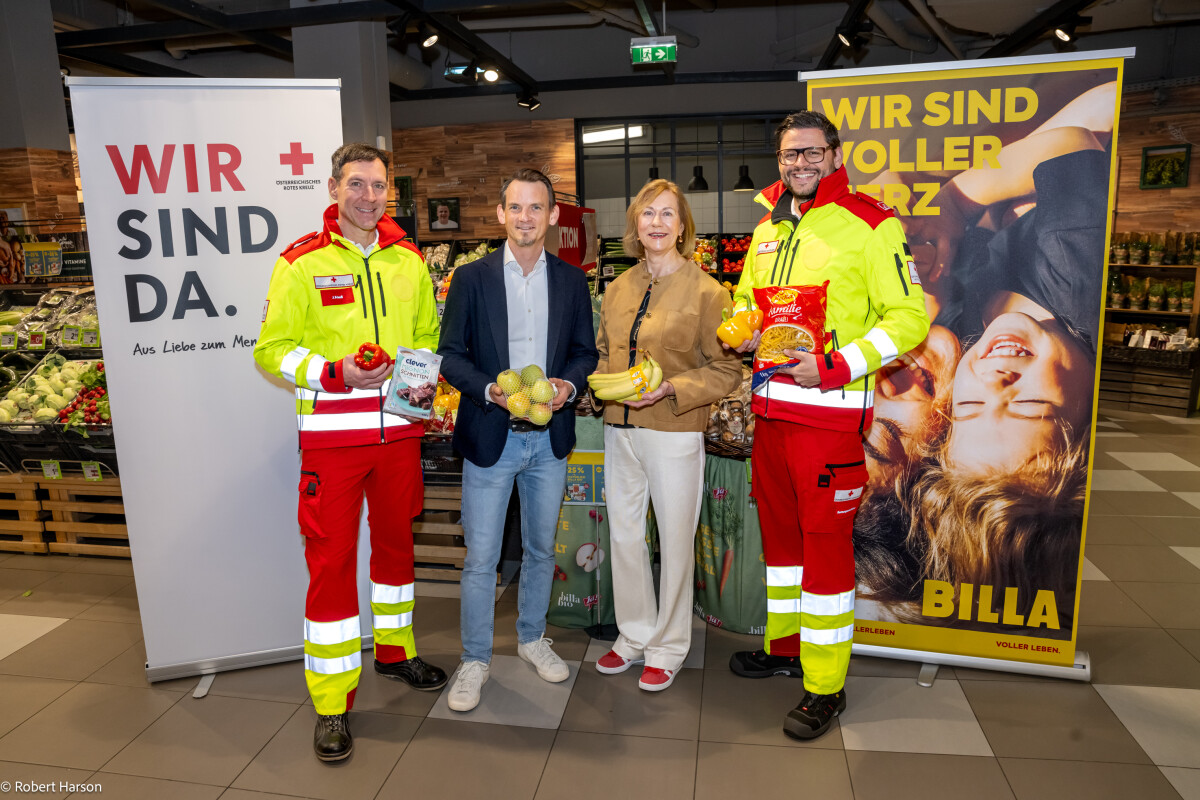 Four people standing in a grocery store, with two wearing emergency service uniforms holding packaged goods and apples. Behind them are promotional banners and a display of fresh produce.