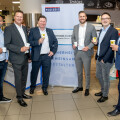 Six men in suits stand together in a public indoor space, holding coffee cups and smiling. A banner behind them reads "POLIZEI" and other text in German. Shelving with food and snacks is visible in the background.