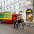 Four people standing in front of a Billa store on a city street, with a truck displaying an advertisement about electric driving next to them. The building has ornate architectural details.