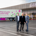 Three men in suits stand in front of a BIPA store and a pink truck with green floral graphics and German text, "Pink ist grün unterwegs. Auch mit diesem E-Lkw." The truck emphasizes sustainable transportation.