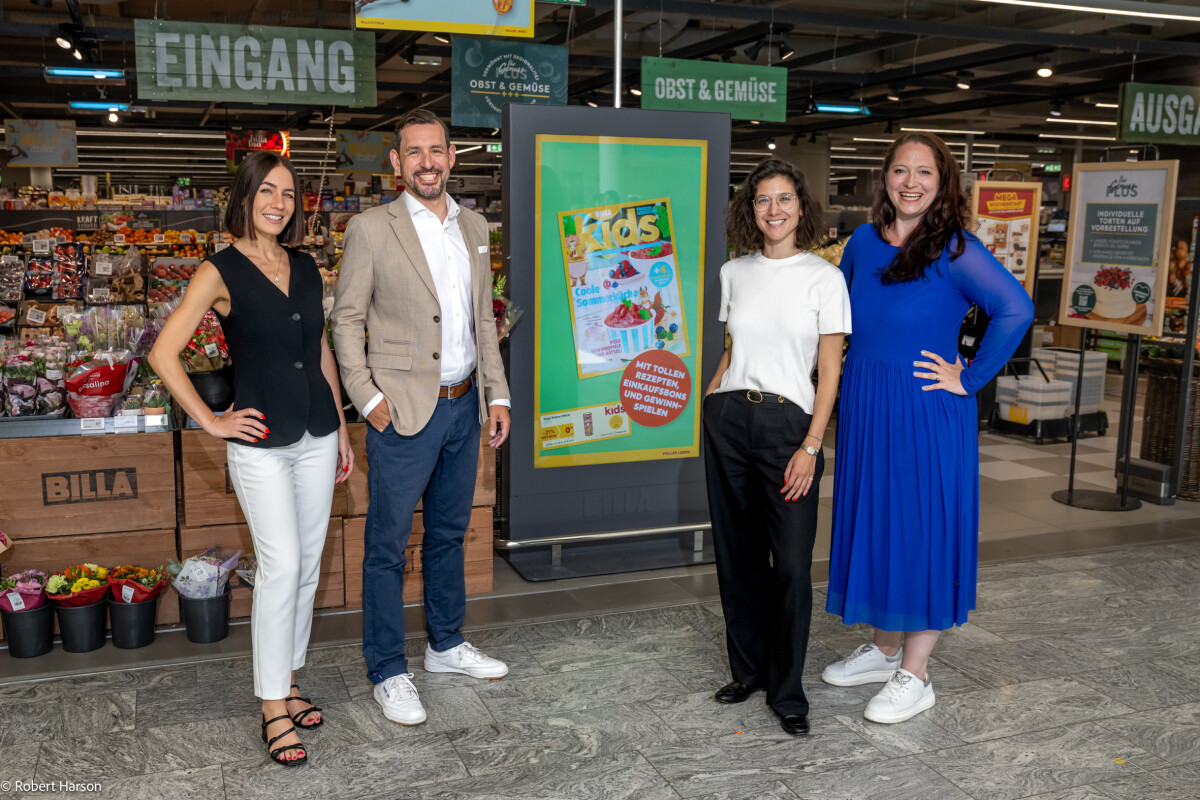 Four people standing and smiling in a grocery store, with a digital display showing a colorful "Kids" advertisement in the background. The store is well-lit and features produce sections, with signage in German.
