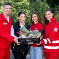 Four people are standing outdoors, holding a box filled with assorted vegetables and groceries. Two of them are wearing red jackets with Red Cross badges, and the others are in casual clothes. Lush greenery is visible in the background.
