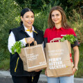 Two women are standing outdoors holding paper bags with leafy greens, wearing branded shirts from Billa and Penny. They are smiling and standing on a pathway with greenery in the background.