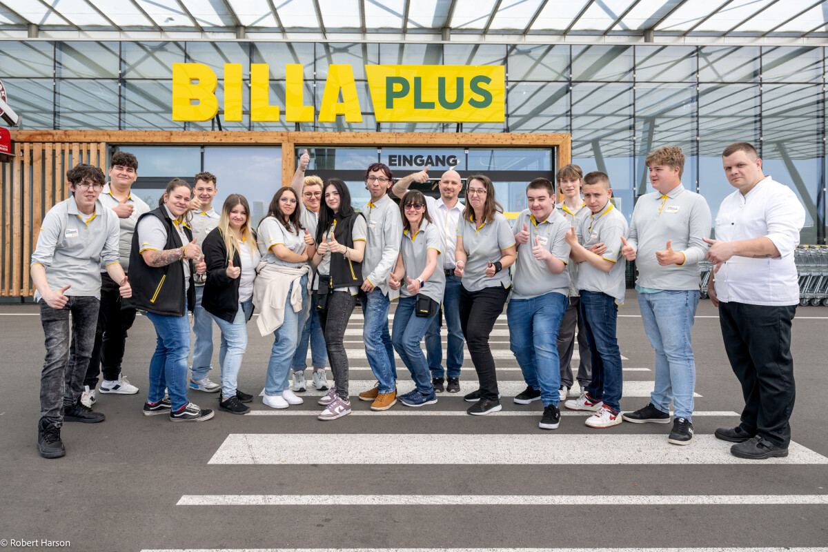 A group of employees in uniform stand in front of a BILLA PLUS store entrance. They are smiling and giving thumbs-up gestures. The building has a modern, glass-front design.