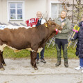 Three people standing with a brown and white cow marked "45." One person holds a sign with a red "ja!" and another holds a yellow "65" sign. All are smiling, and the cow has a red ribbon on its head. A wooden fence and building are in the background.