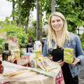 A woman smiling while standing by a table filled with various organic products, including juices, snacks, and olive oil. The setting is a lush garden with greenery in the background.