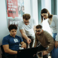 Four people are gathered around a laptop, engaged in discussion. One person is seated while the others stand, with one pointing at the screen. A poster on the wall reads, "Let's shape the future of retail together."