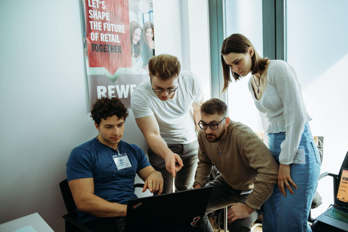 Four people are gathered around a laptop, engaged in discussion. One person is seated while the others stand, with one pointing at the screen. A poster on the wall reads, "Let's shape the future of retail together."