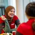 A woman wearing a red and green uniform and a brown hijab is smiling at a colleague during a break. They appear to be sitting at a table with food and drinks.