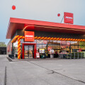A modern store facade with bright red and yellow balloon decorations around the entrance. Red signs display the name "PENNY" above the door. Balloons and pallets of goods are seen outside. The sky is clear, with some large red balloons floating above.