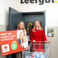 Two women stand in front of a door with a "Leergut" sign. One holds a sign about helping families in need in Austria, featuring the Austrian Red Cross logo. The other woman stands beside a shopping cart filled with empty plastic bottles.