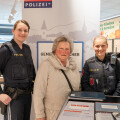 Three women smiling indoors at a grocery store. Two of them are in police uniforms, and the third is an older woman wearing a beige jacket. A sign behind them has the word "Polizei."