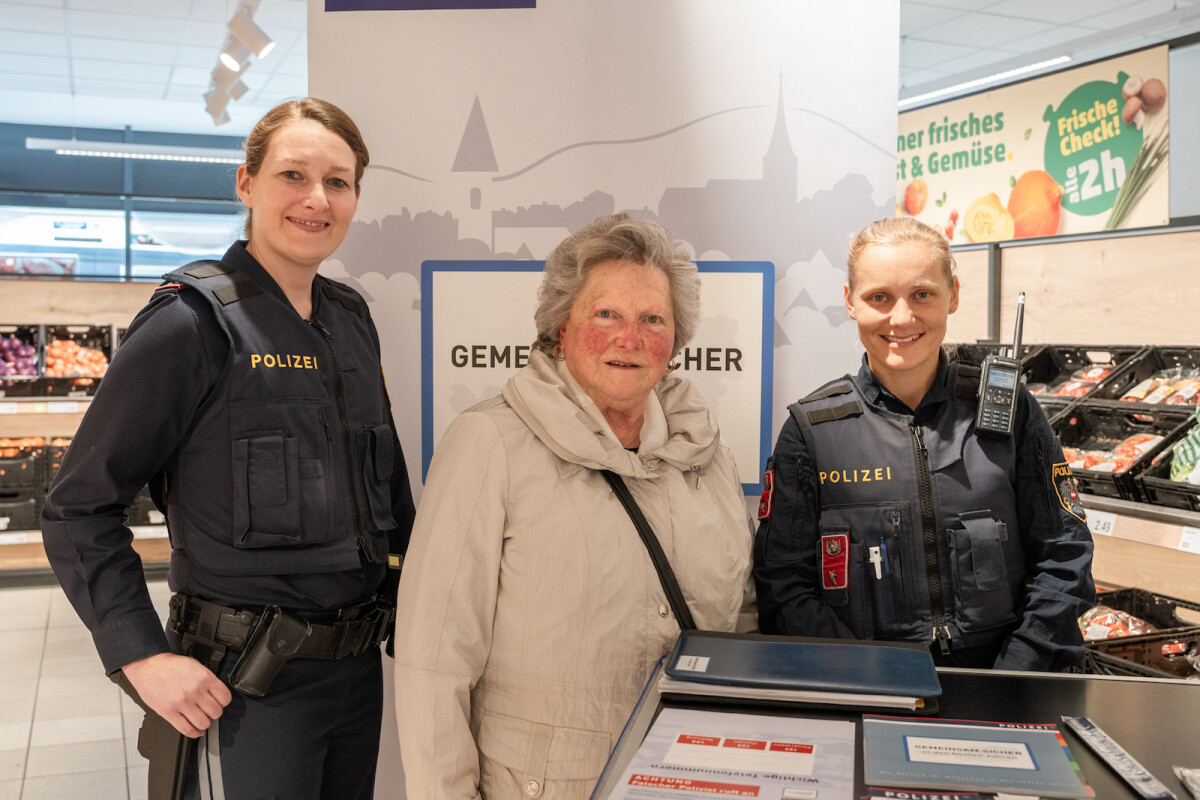 Three women smiling indoors at a grocery store. Two of them are in police uniforms, and the third is an older woman wearing a beige jacket. A sign behind them has the word "Polizei."
