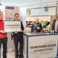 Four people standing in a supermarket setting, with two in police uniforms. They are next to a display for "GEMEINSAM.SICHER" with REWE International AG, featuring a country map outline and a police banner. One person holds a sign with the same text.
