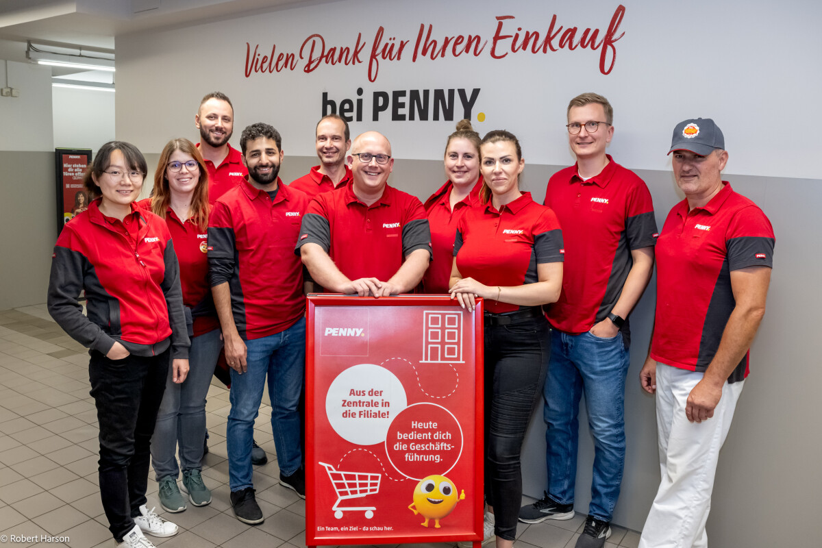 A group of people in matching PENNY uniforms smiling and posing for a photo indoors. They are standing near a sign with text and graphics, against a wall with a thank you message.