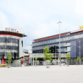 Two modern office buildings featuring glass facades, one labeled "REWE" and the other "BILLA," with a spacious parking area in the foreground and a cloudy sky above.