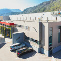 Large gray industrial building with "Rewe Group" signage, set against a mountainous backdrop. A red delivery truck and two cars are parked nearby. Clear blue sky above.