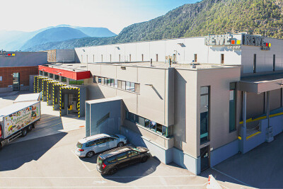 Large gray industrial building with "Rewe Group" signage, set against a mountainous backdrop. A red delivery truck and two cars are parked nearby. Clear blue sky above.