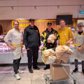 Five men stand in a butcher shop, with two wearing uniforms and three in casual attire, surrounding a shopping cart filled with large pieces of meat. The backdrop features a display of various meats.