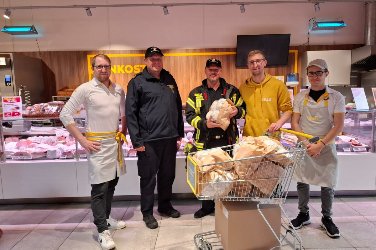 Five men stand in a butcher shop, with two wearing uniforms and three in casual attire, surrounding a shopping cart filled with large pieces of meat. The backdrop features a display of various meats.
