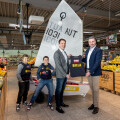 Two men, one holding a sailing vest, pose with two boys in sports jerseys in a supermarket. A small sailboat is displayed behind them in the produce section.