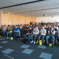 A large group of people seated in a conference room, listening attentively. Some are clapping and others are holding yellow bags. Two people in yellow shirts stand near the entrance, also applauding. The room has wooden walls and a ceiling-mounted projector.