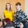 A man and woman stand among a seated group in a classroom setting. The man wears a black and white patterned sweater and a crossbody bag, while the woman wears a bright yellow shirt. Both are focused on something out of frame.