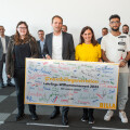 A group of people standing indoors, holding a large sign with multiple signatures and the text "#nextbillageneration Lehrlings-Willkommensevent 2023." A few individuals are positioned at the front, and a row of people is visible in the background.