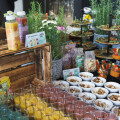 A colorful display of bottled juices, fresh salads, and small bowls of dessert with nuts, arranged on a wooden crate and tiered stands. Decorated with potted herbs and a variety of plants and flowers.
