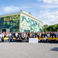 A large group of people posing in front of a supermarket with a colorful mural depicting fruits and vegetables. Some are holding yellow and orange signs, and others have a large poster. The sky is blue with a few clouds.