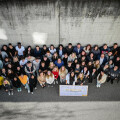 A large group of people stand together on a pavement next to a concrete wall. They are looking up at the camera, with some wearing yellow shirts. A colorful sign is held at the front.