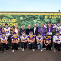 A group of American football players in purple and white uniforms pose with several adults in casual attire. They are standing outside in front of a building with "BILLA Plus" signage and two yellow banners. The players are wearing helmets and jerseys with numbers.