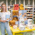 A woman in a white blouse and jeans stands in a grocery store holding a food product box. Behind her, there is a yellow table stacked with various packaged food items. Shelves filled with more products are visible in the background.