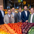 Six men in business attire stand smiling behind a display of assorted fresh vegetables, including tomatoes, carrots, red onions, and pumpkins, inside a grocery store.