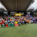 A large group of children and adults in colorful sports uniforms are gathered indoors on a turf field, posing for a group photo. Some hold banners and trophies. The ceiling has skylights, and the walls display various sponsor banners.