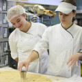 Two bakers in white uniforms and head coverings cut dough into heart shapes on a large work surface in a bakery kitchen. Shelves and baking equipment are visible in the background.