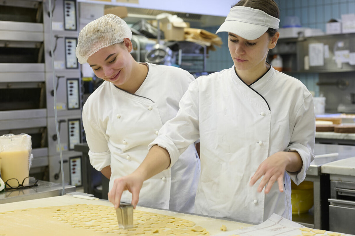 Two bakers in white uniforms and head coverings cut dough into heart shapes on a large work surface in a bakery kitchen. Shelves and baking equipment are visible in the background.
