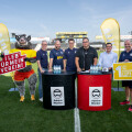 A group of men standing on a soccer field next to branded tables with drinks. A person in a boar costume holds a sign. Two large banners with logos are displayed behind them. Stadium seats and a scoreboard are visible in the background.