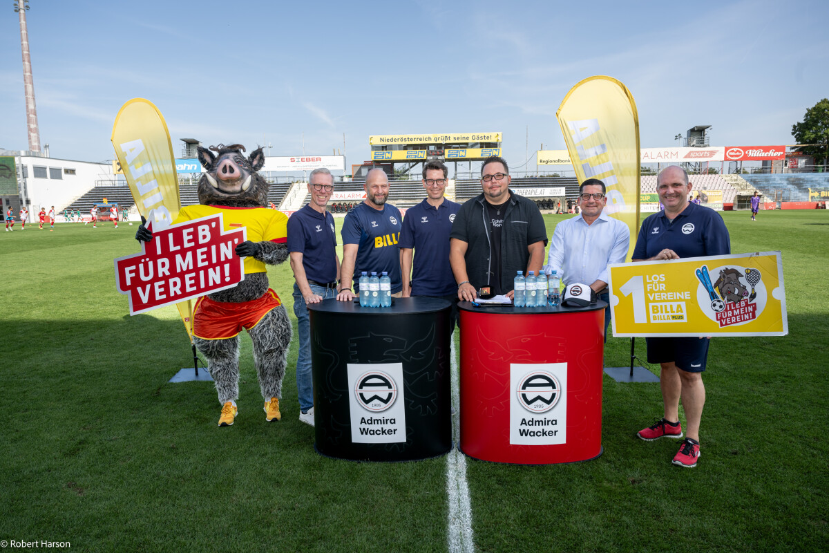 A group of men standing on a soccer field next to branded tables with drinks. A person in a boar costume holds a sign. Two large banners with logos are displayed behind them. Stadium seats and a scoreboard are visible in the background.