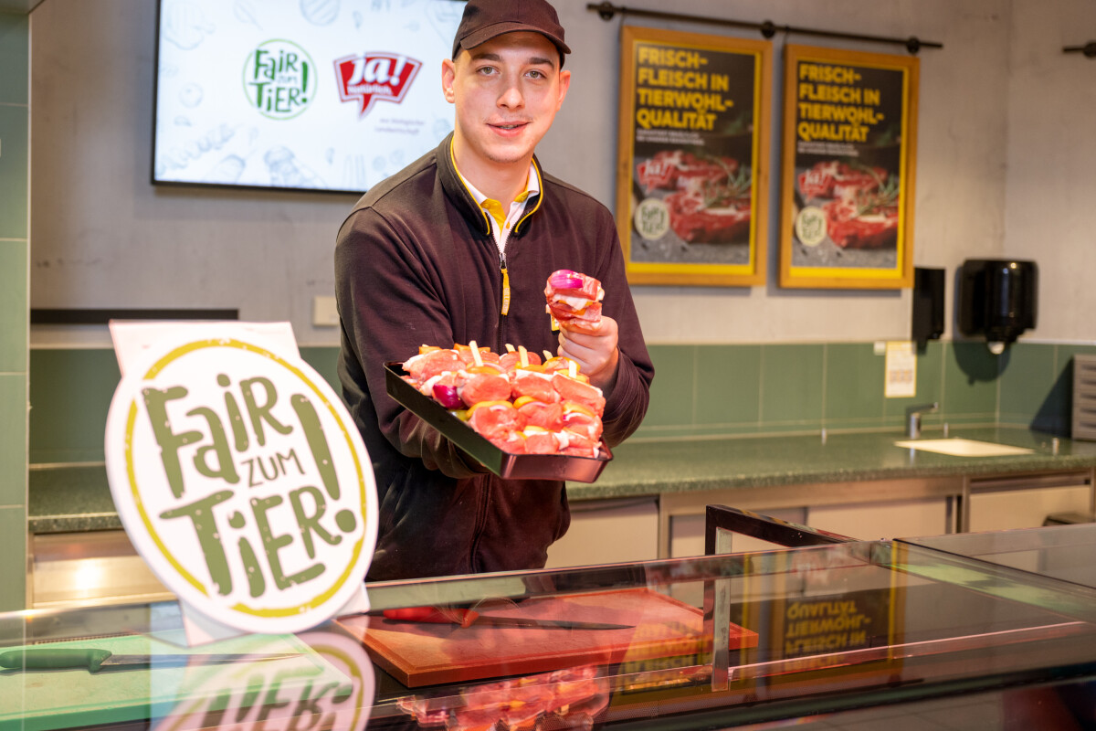 A man in a black cap and uniform, holding a tray of raw meat cuts, stands behind a counter in a meat shop. The shop features green tiles and posters promoting quality and animal welfare.