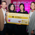 Four people smiling and holding a large check for €8,500 from Billa Plus, in front of a backdrop with sponsor logos.