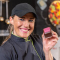 A smiling woman in a black cap and chef's uniform holds a chocolate with a pink label. Cooking utensils and blurred colorful decorations are in the background.
