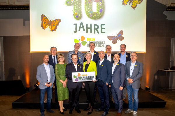 A group of people standing on a stage, smiling and posing for a photo. They are holding a sign with a colorful logo. The backdrop displays butterflies and the text "10 Jahre." The setting appears to be a formal event or celebration.