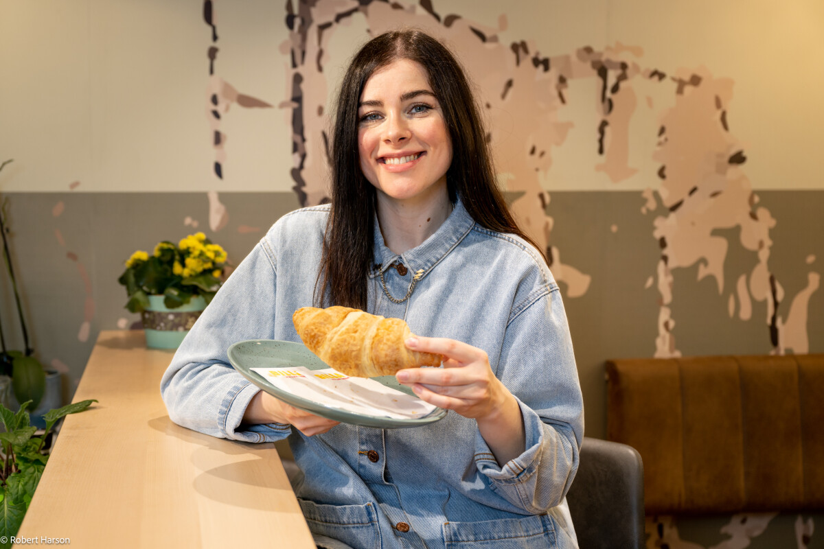 A woman with long dark hair is smiling and holding a plate with a croissant. She is wearing a light denim jacket and sitting at a table in a cafe. Yellow flowers and a textured wall are in the background.