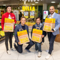 Four people smiling and holding shopping bags stand in front of a BILLA store entrance surrounded by fresh produce displays, including oranges and apples. The environment is bright and inviting.
